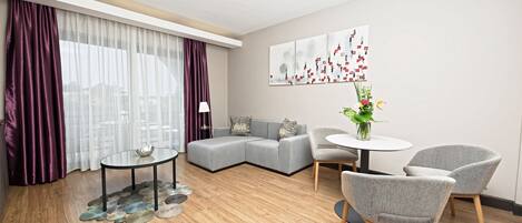 A modern living room with a grey sectional sofa, a round glass coffee table, two grey armchairs, and a small round table with a vase of flowers.