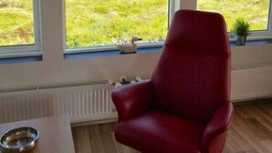 A room with a red chair, a small table with a bowl, and large windows offering a view of a green field and distant mountains. - Blue View Cabin 1A with Hot Tub (Bláskógabyggd)