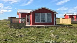 A red single-story house with a large window and a door, surrounded by a grassy field and a wooden fence. - Blue View Cabin 1A with Hot Tub (Bláskógabyggd)