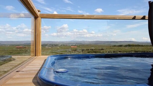 A hot tub on a wooden deck with a view of a grassy landscape and distant hills. - Blue View Cabin 1A with Hot Tub (Bláskógabyggd)