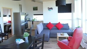 A modern living room with a grey sofa, red and white cushions, a red chair, a dining table with chairs, and a kitchen area in the background. - Blue View Cabin 1A with Hot Tub (Bláskógabyggd)