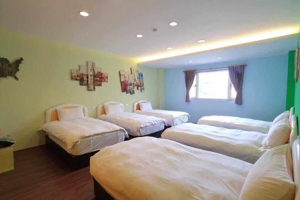A hotel room with four beds arranged in a row, a window with curtains, and artwork on the walls.