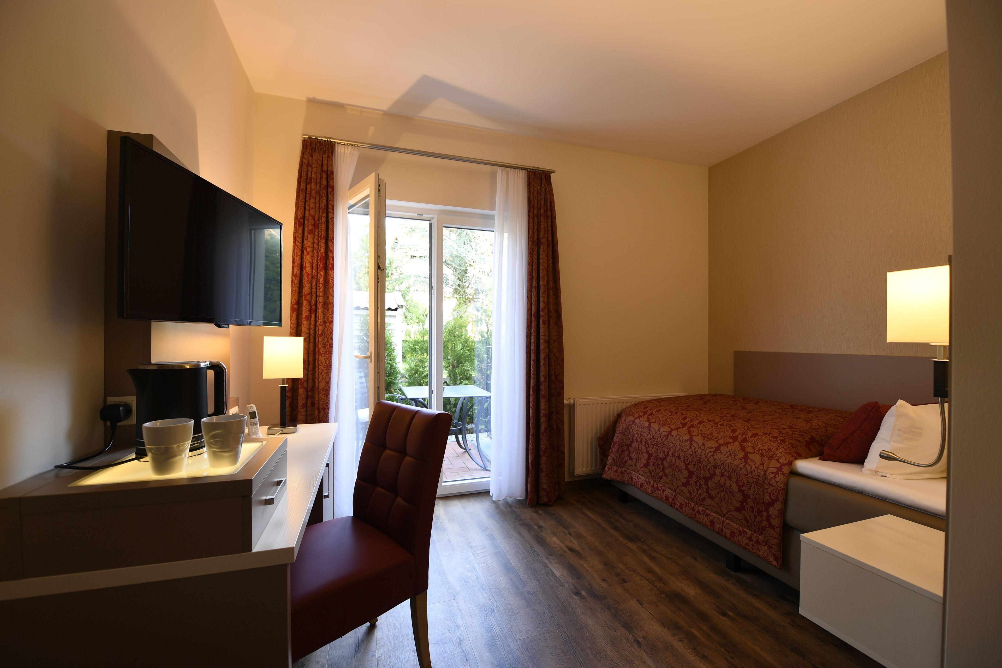A hotel room with a bed, a desk with a coffee maker, a chair, a TV, and a window with curtains.