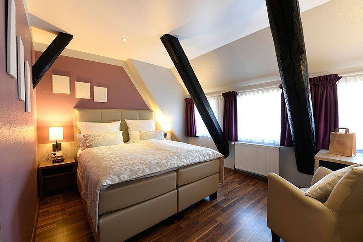 A modern hotel room with a bed, a nightstand, a chair, and a window with curtains.