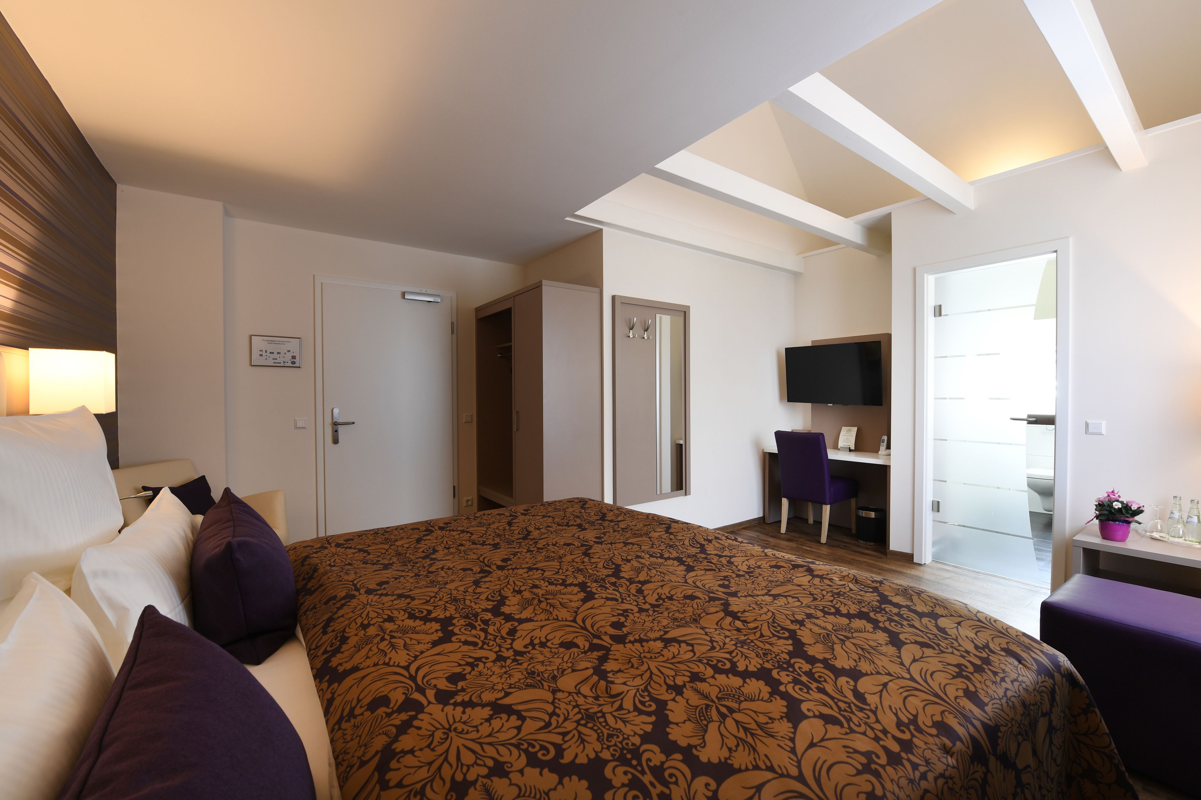 Habitación de hotel con una cama grande, un escritorio, una silla, un televisor y una puerta que da a otra habitación.