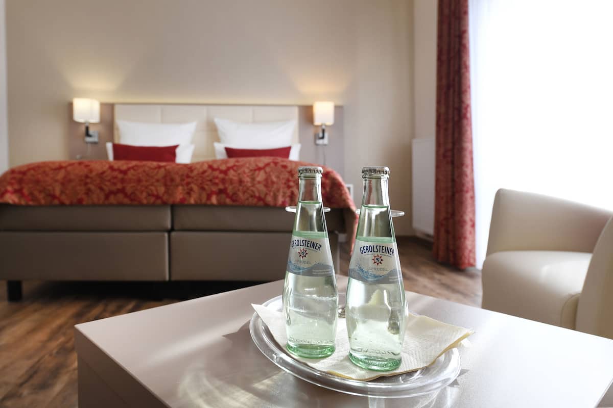 A hotel room with a bed, two bottles of Gerolsteiner water, a white table, and a white chair.