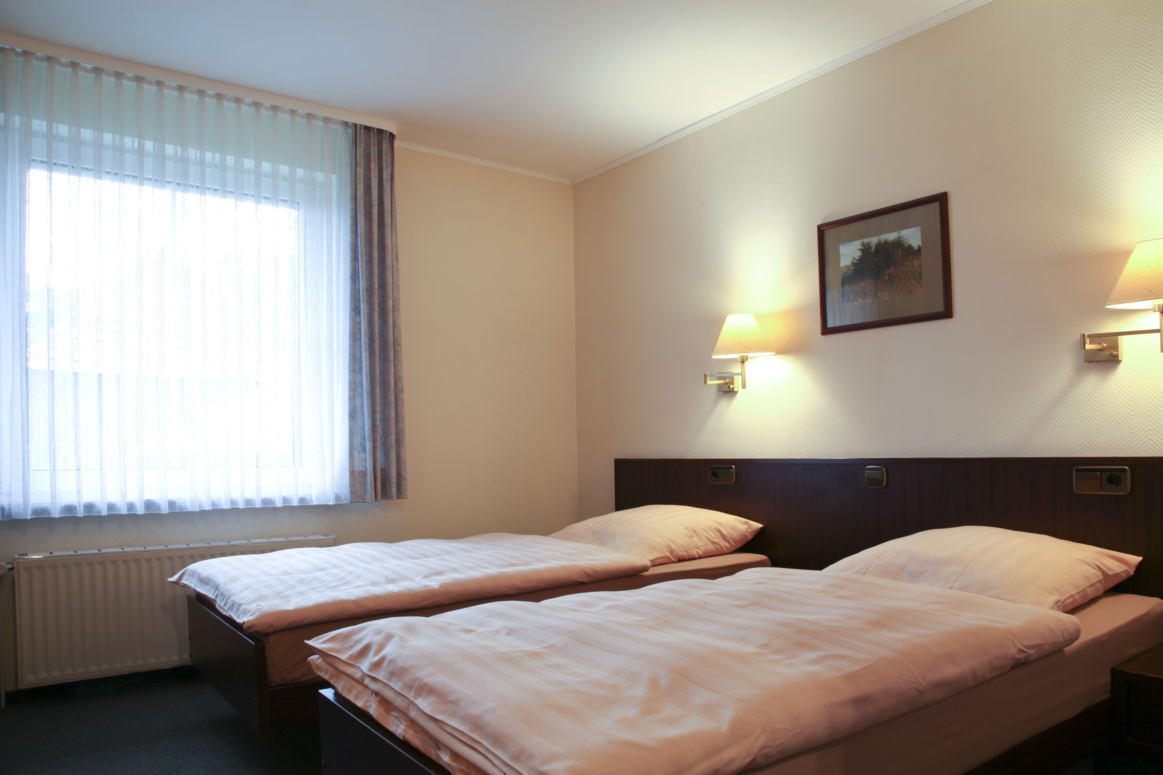 Quarto de hotel com duas camas, uma janela com cortinas finas, um radiador e luminárias fixadas na parede.