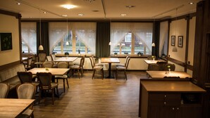 A hotel dining area with multiple tables and chairs, large windows with curtains, and a view of a building outside. - Landgasthof Voltmer (Burgdorf)