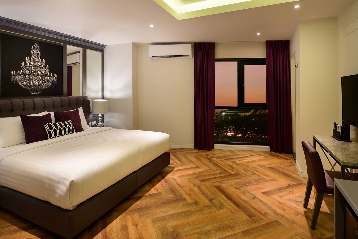 A hotel room with a large bed, a chandelier, a dining area, and a view of the city at sunset.