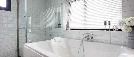 A modern bathroom with a glass-enclosed shower, a large white bathtub, and a window with blinds.