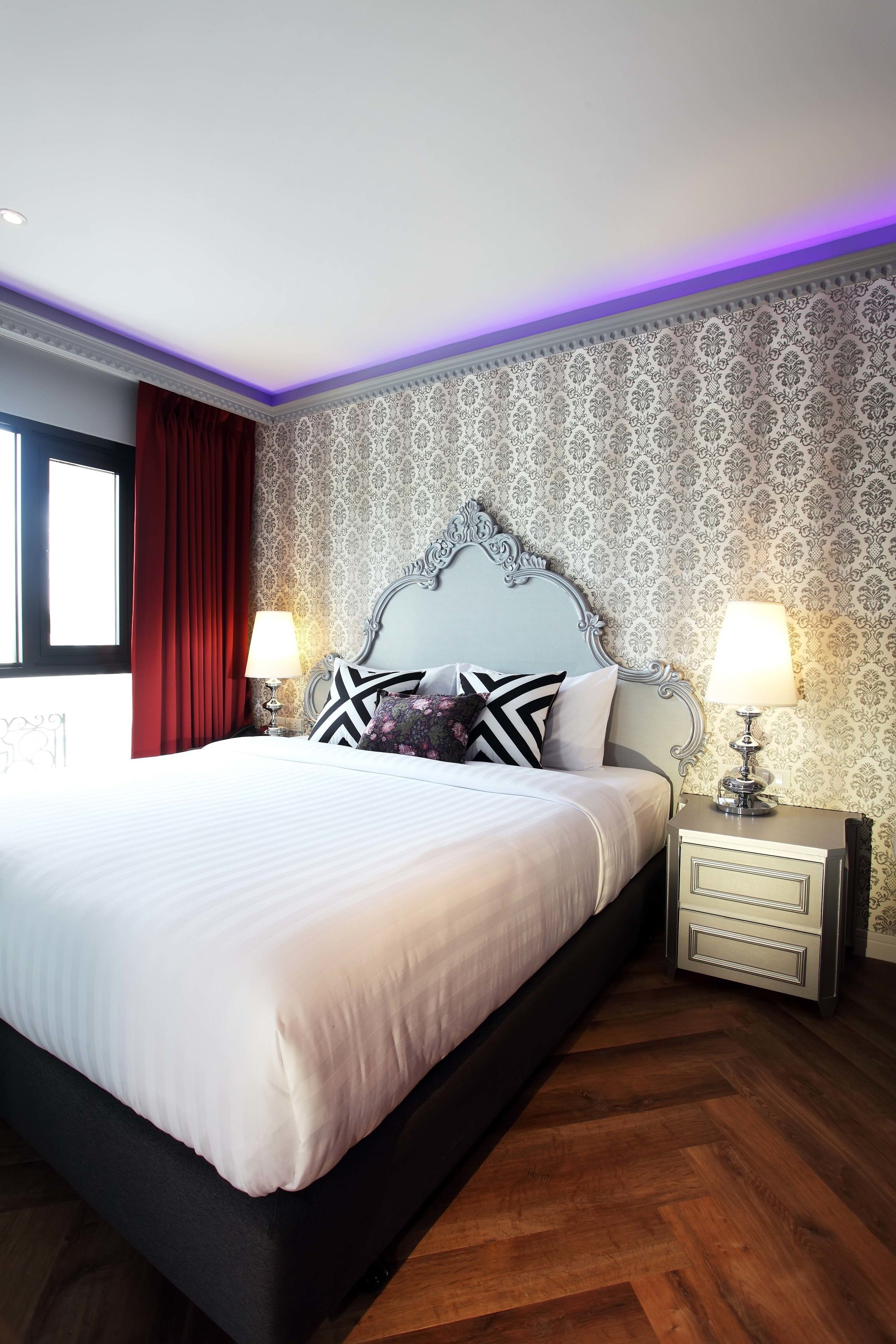 A neatly made bed with a white duvet and black and white pillows, flanked by two bedside tables with lamps, in a room with patterned wallpaper and wooden flooring.