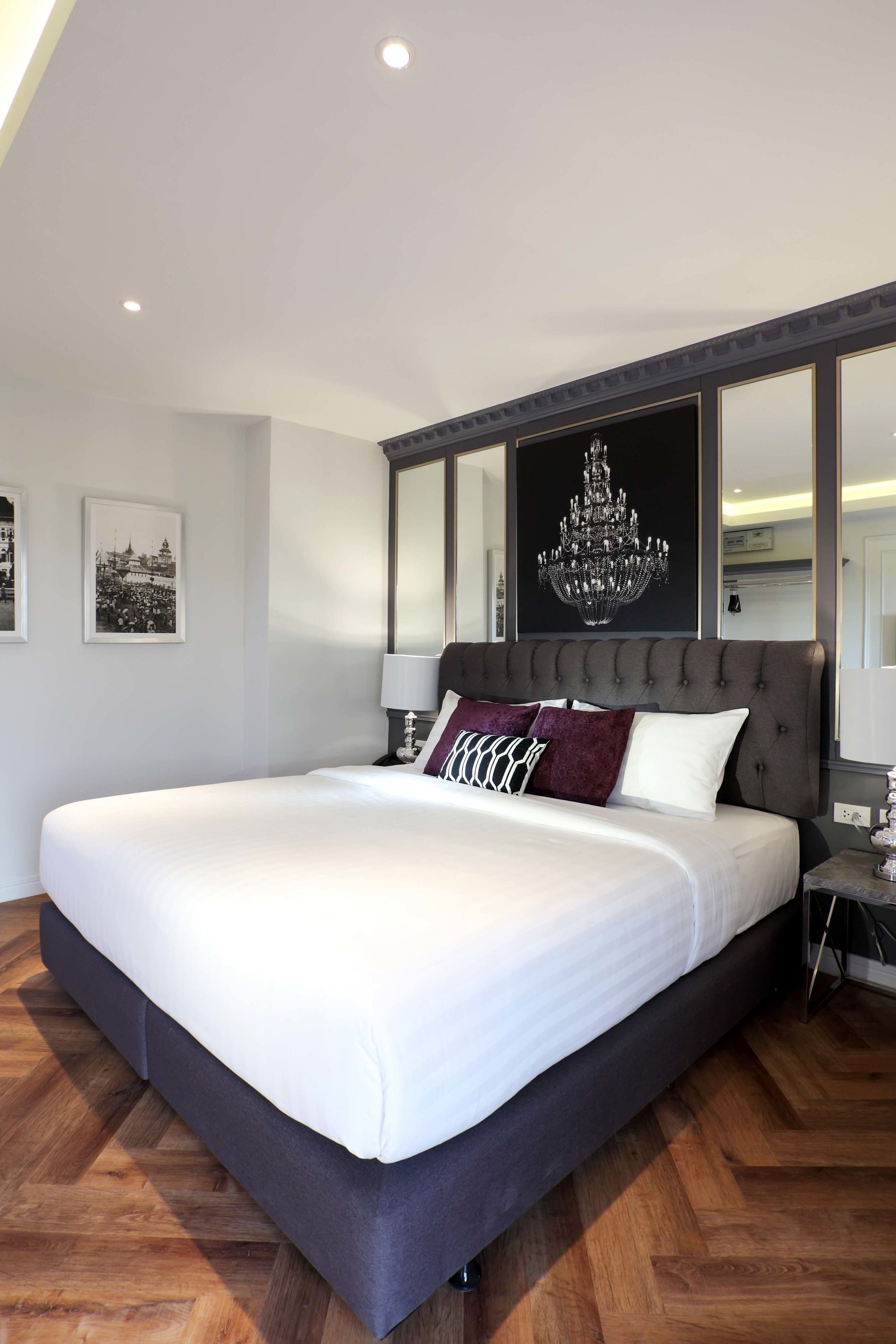A large bed with a dark headboard, white bedding, and multiple pillows. There are framed pictures on the wall and a chandelier above the bed.