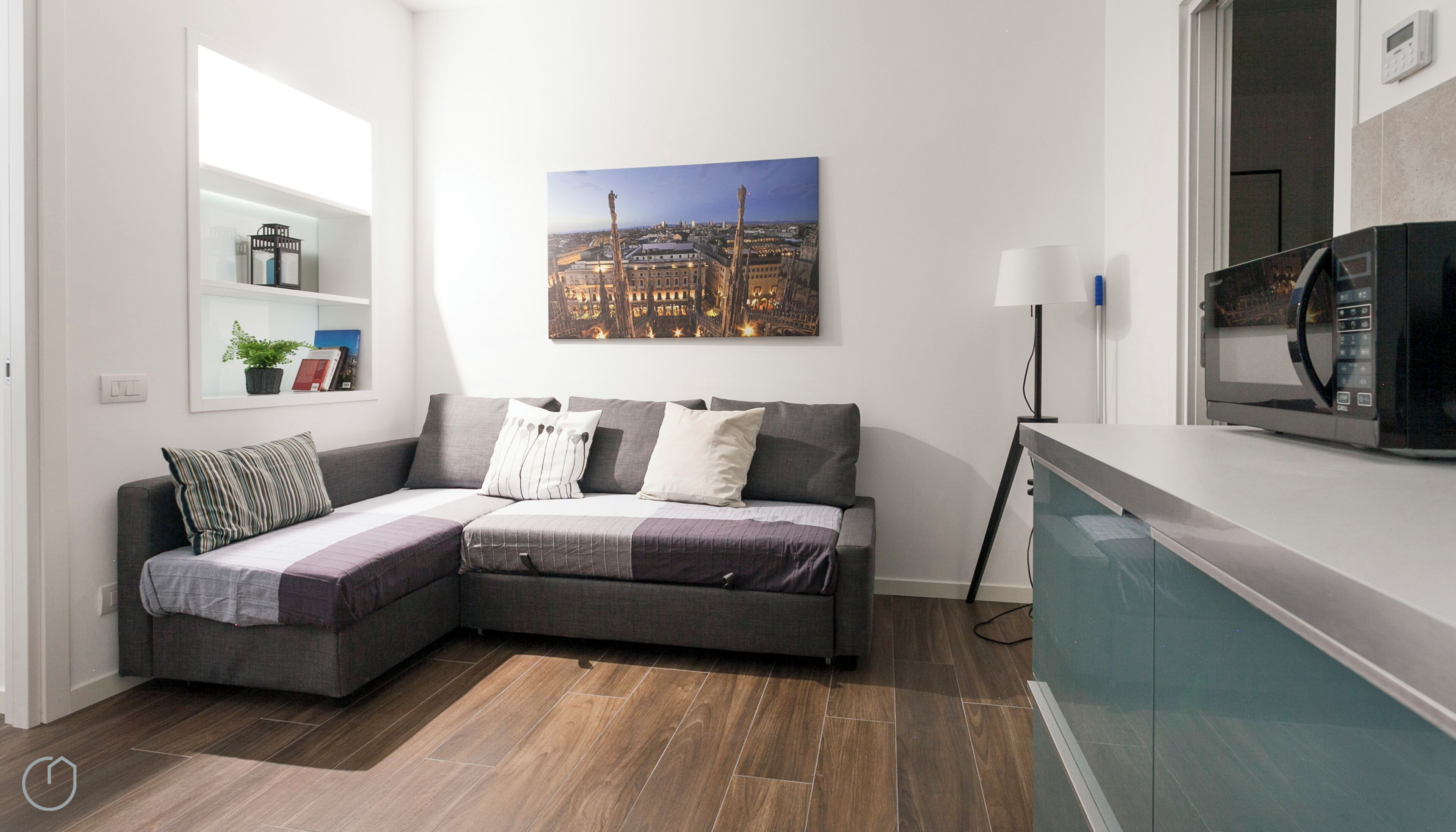 Ein modernes Wohnzimmer mit einem grauen Ecksofa, einem Stadtbild-Gemälde, einer kleinen Ablage mit Büchern und einer Pflanze, einer Stehlampe und einer Mikrowelle auf einer Arbeitsfläche.