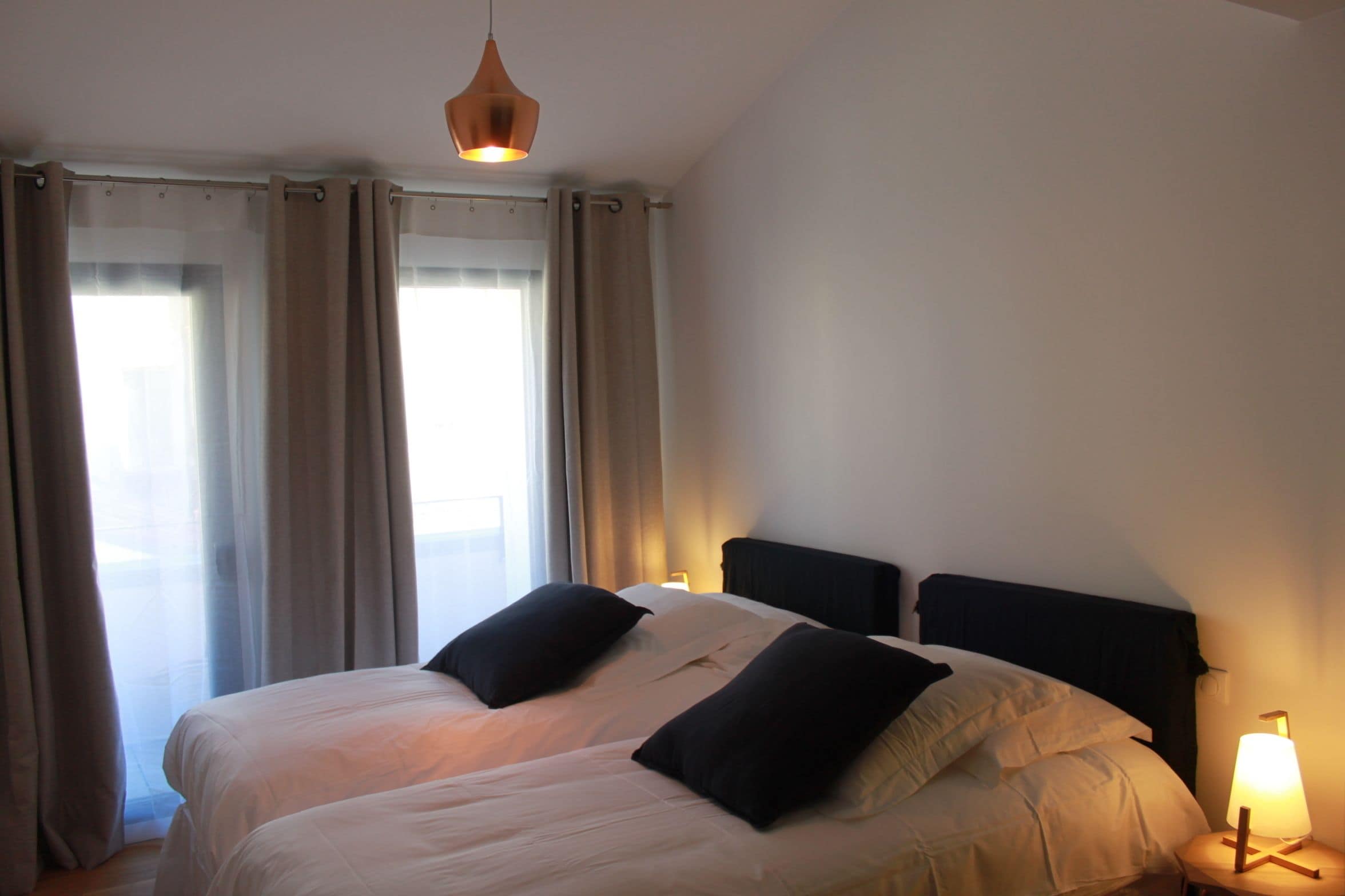 Ein Doppelbett mit zwei schwarzen Kissen, eine hängende Pendelleuchte und ein Nachttisch mit Lampe.