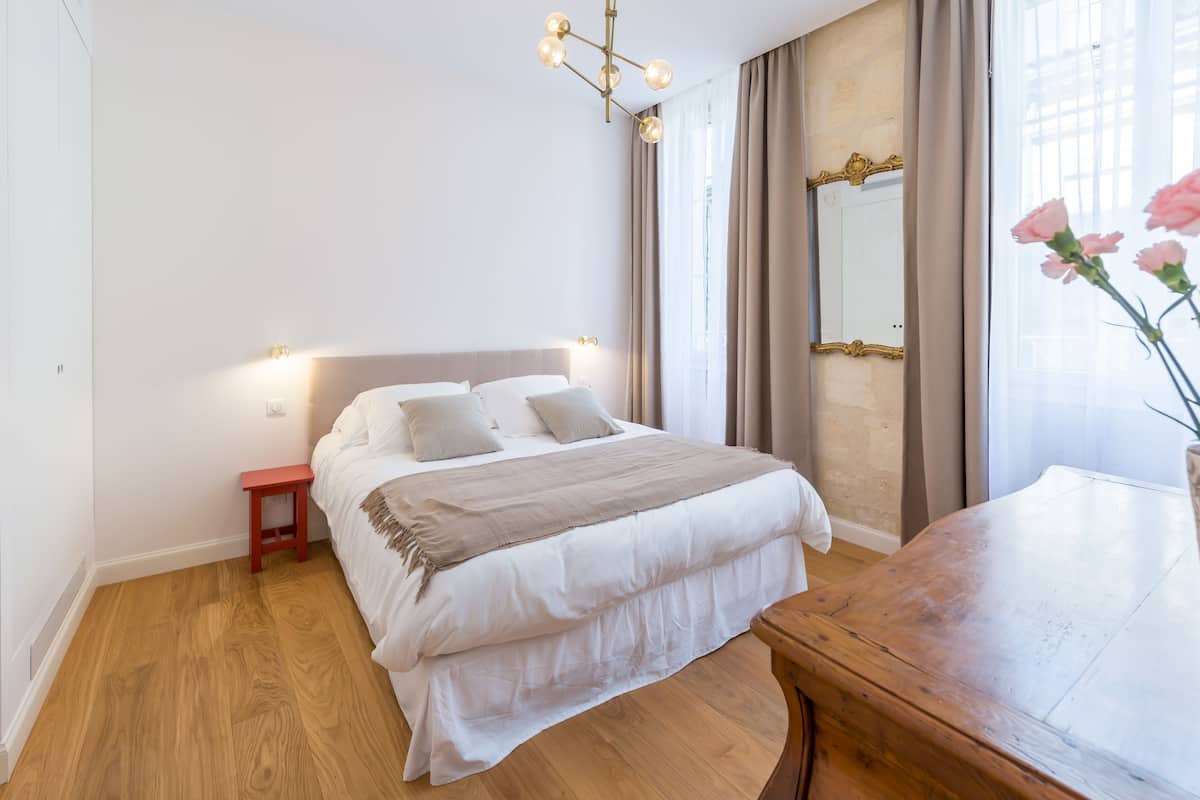 Un dormitorio ordenado con suelo de madera, una cama grande con ropa blanca, una mesita de noche y un espejo con cortinas.