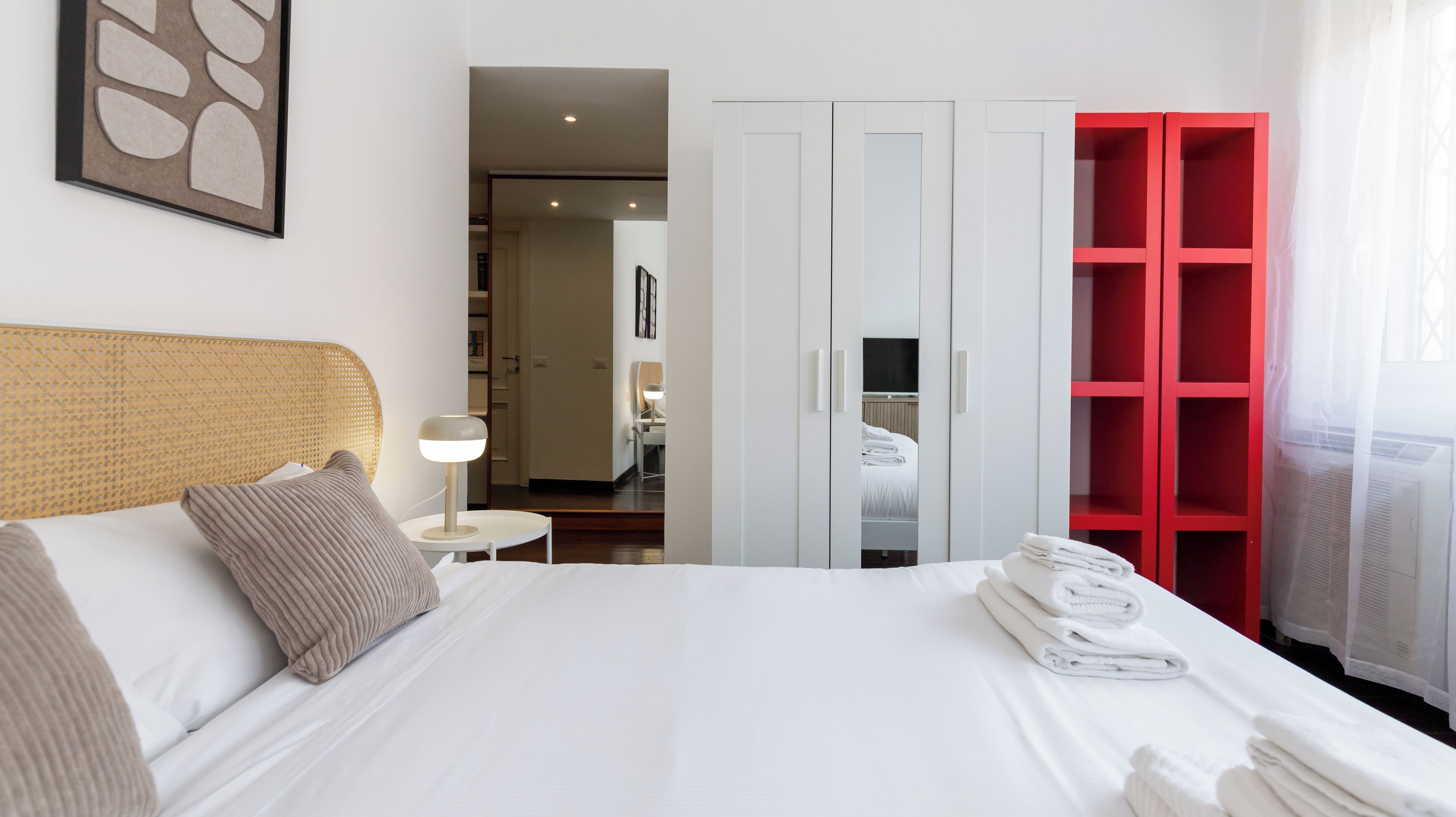 A hotel room with a bed, a nightstand, a red wardrobe, and a white door leading to a bathroom.