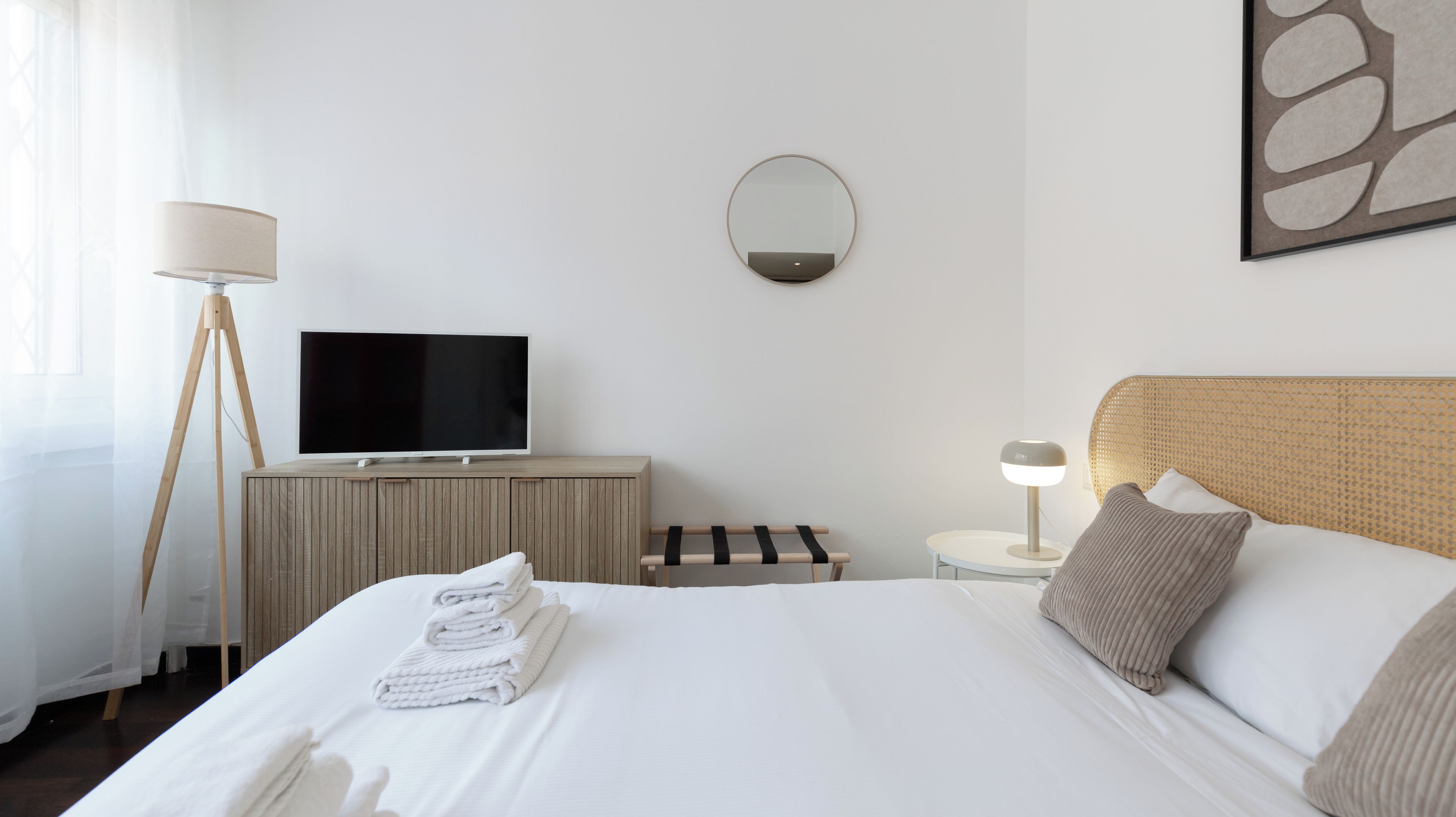 Habitación de hotel con cama, televisión, mesita de noche con lámpara, espejo en la pared y ventana con cortinas.