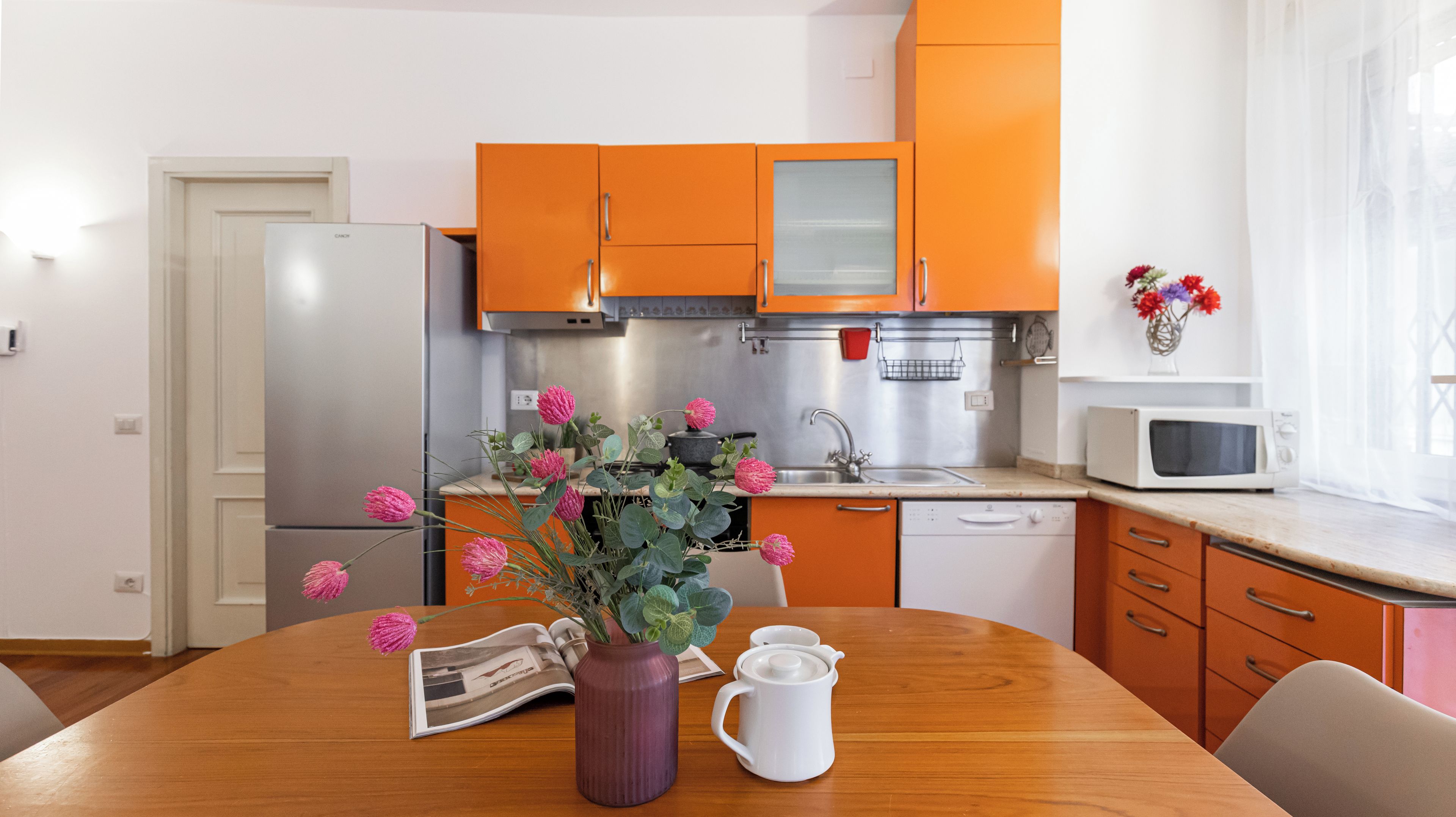 A modern kitchen with orange cabinets, stainless steel appliances, and a wooden dining table with a vase of flowers.