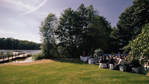 An outdoor wedding ceremony with guests seated on white-covered tables, a wooden dock, and a lake in the background. - Zum Rittmeister (Werder (Havel))