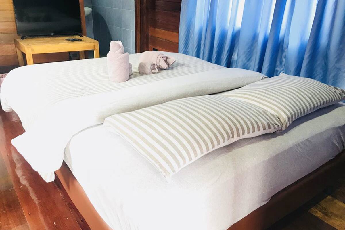 A double bed with white linens and striped pillows, a wooden headboard, a small wooden table with a television, and a blue curtain.