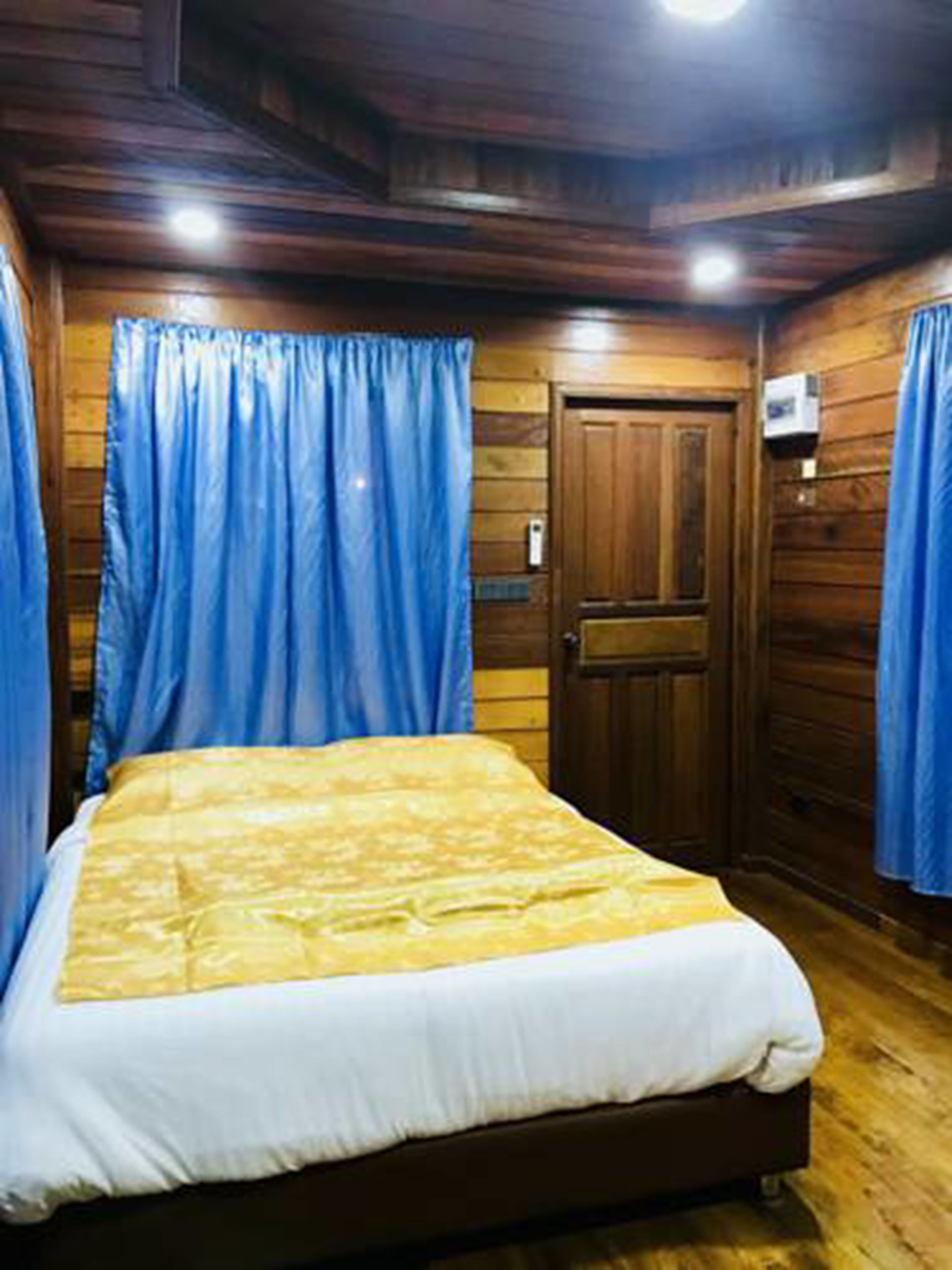 A bedroom with a wooden floor, a bed with a white and yellow bedspread, and blue curtains.