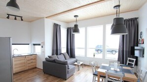 A modern apartment with a kitchen, dining area, and living room. It features a grey sofa, a dining table with chairs, and a TV mounted on the wall. - Blue View Cabin 4A with Hot Tub (Bláskógabyggd)