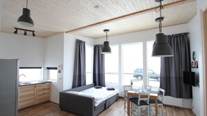 A modern, compact room with a bed, a small table, and a chair. It features a kitchen area with a refrigerator and cabinets, large windows with curtains, and a ceiling with wooden beams. - Blue View Cabin 4A with Hot Tub (Bláskógabyggd)