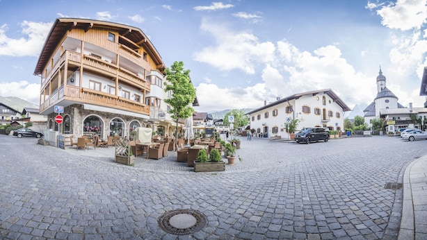 Une place pavée, avec un bâtiment en bois doté de balcons, et une église coiffée d’un dôme en arrière-plan.
