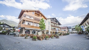 Une place pavée avec des tables en plein air, un paysage montagneux en toile de fond et une architecture européenne traditionnelle.