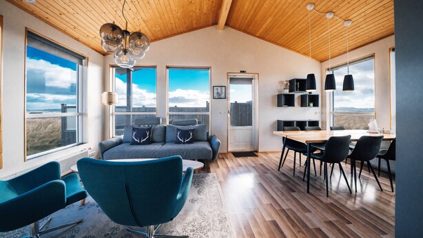 A spacious living area with a wooden ceiling, a gray sofa with decorative pillows, a blue armchair, a dining table with chairs, and large windows offering a view of the sky and landscape. - Blue View Cabin 5A with Hot Tub (Bláskógabyggd)