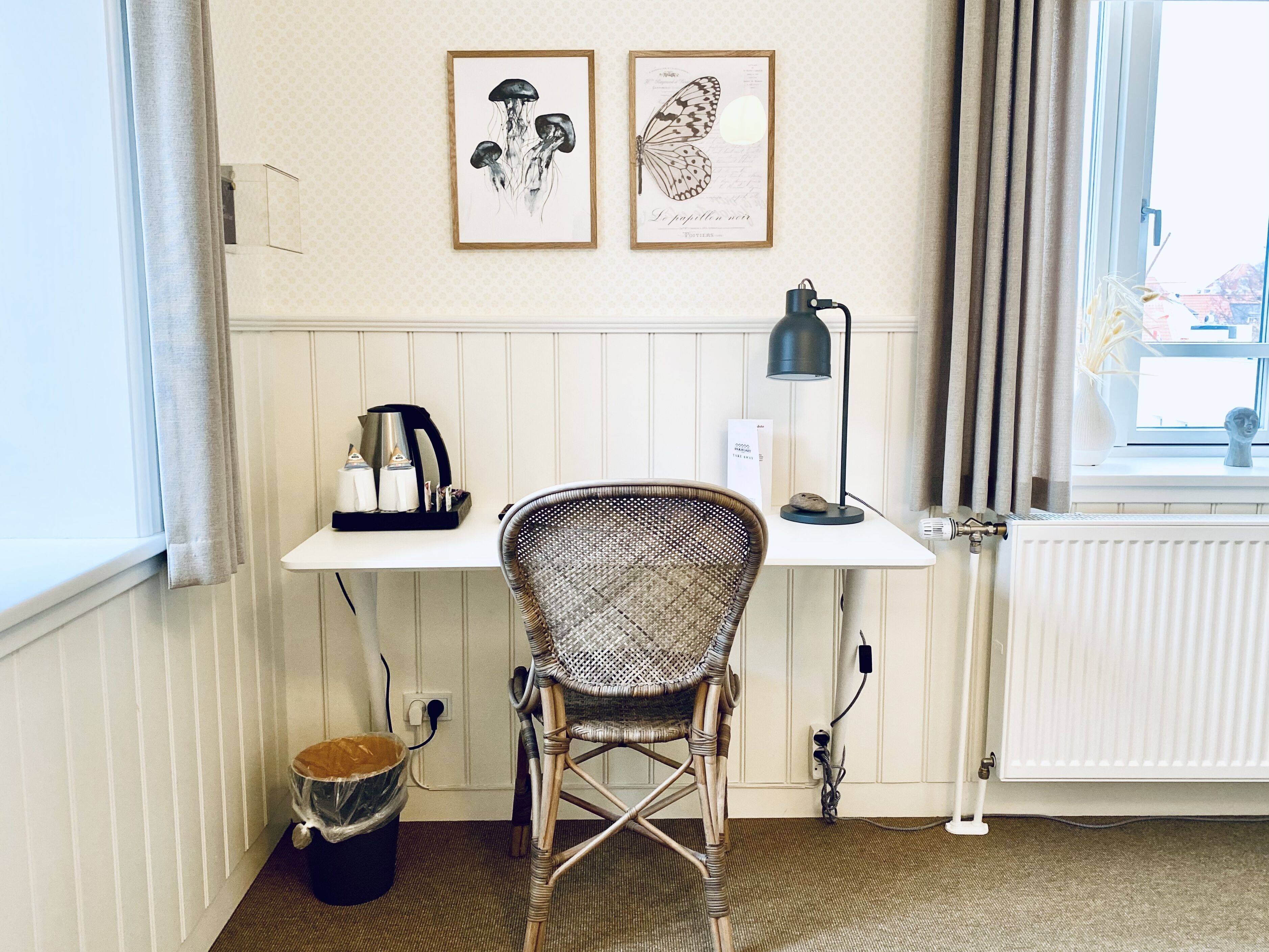 A room with a white desk, a wicker chair, a coffee maker, a framed butterfly picture, and a radiator.