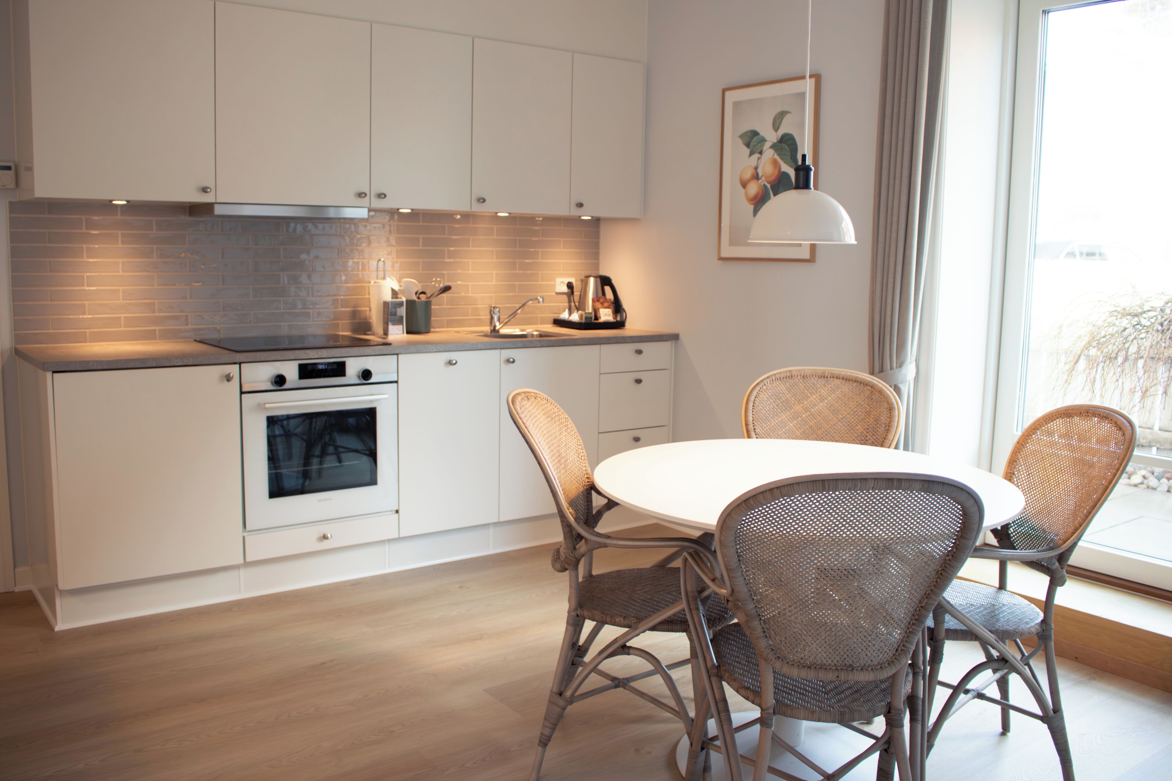 Una cocina moderna con armarios blancos, horno empotrado, una mesa de comedor con sillas de mimbre y un ventanal con vistas.