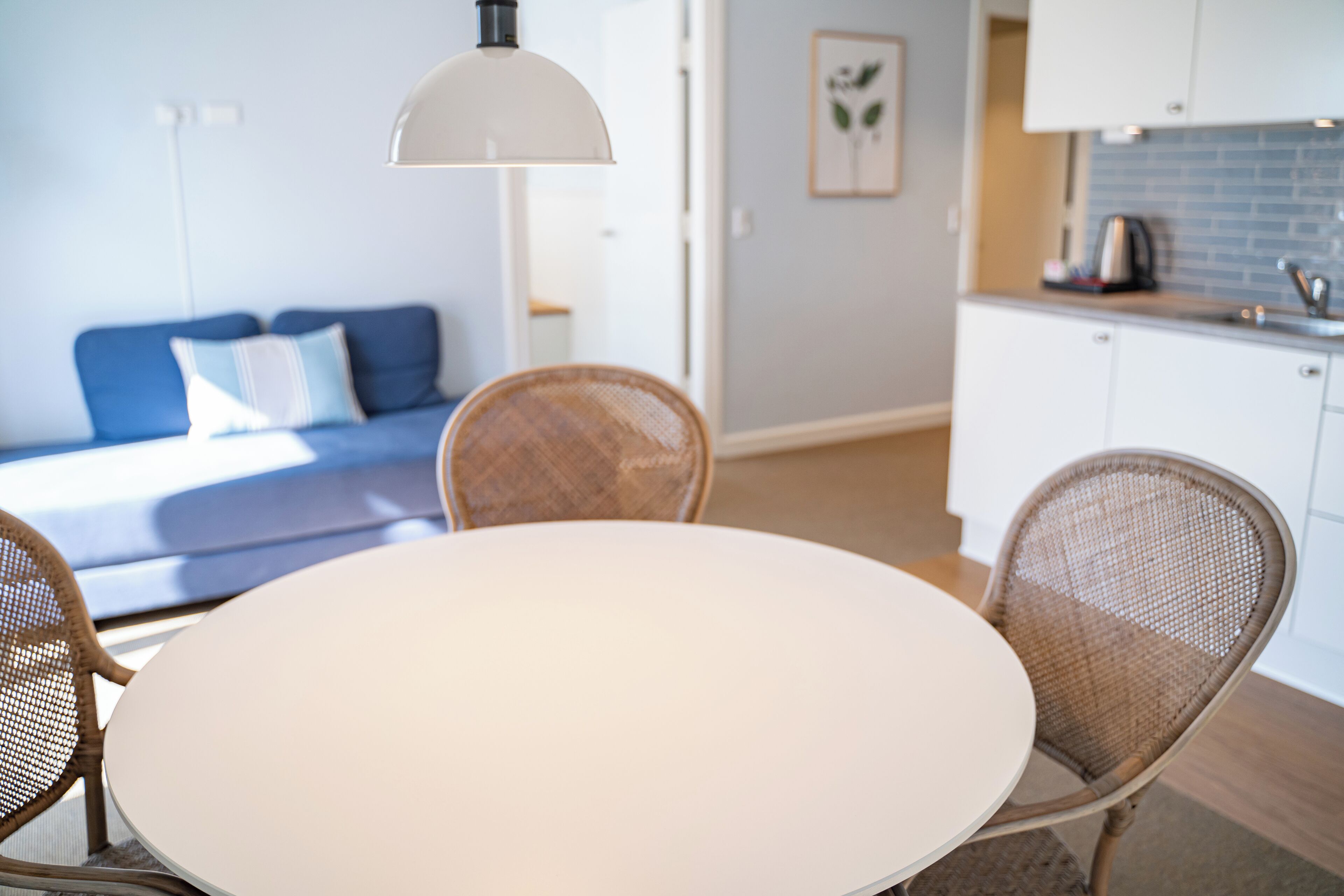 Una habitación moderna con una mesa redonda blanca, sillas de mimbre, un sofá azul con almohadillas blancas, una lámpara de techo blanca y una zona de cocina con armarios blancos y salpicadero azul.