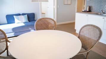 Una habitación moderna con una mesa redonda blanca, sillas de mimbre, un sofá azul con almohadillas blancas, una lámpara de techo blanca y una zona de cocina con armarios blancos y salpicadero azul.