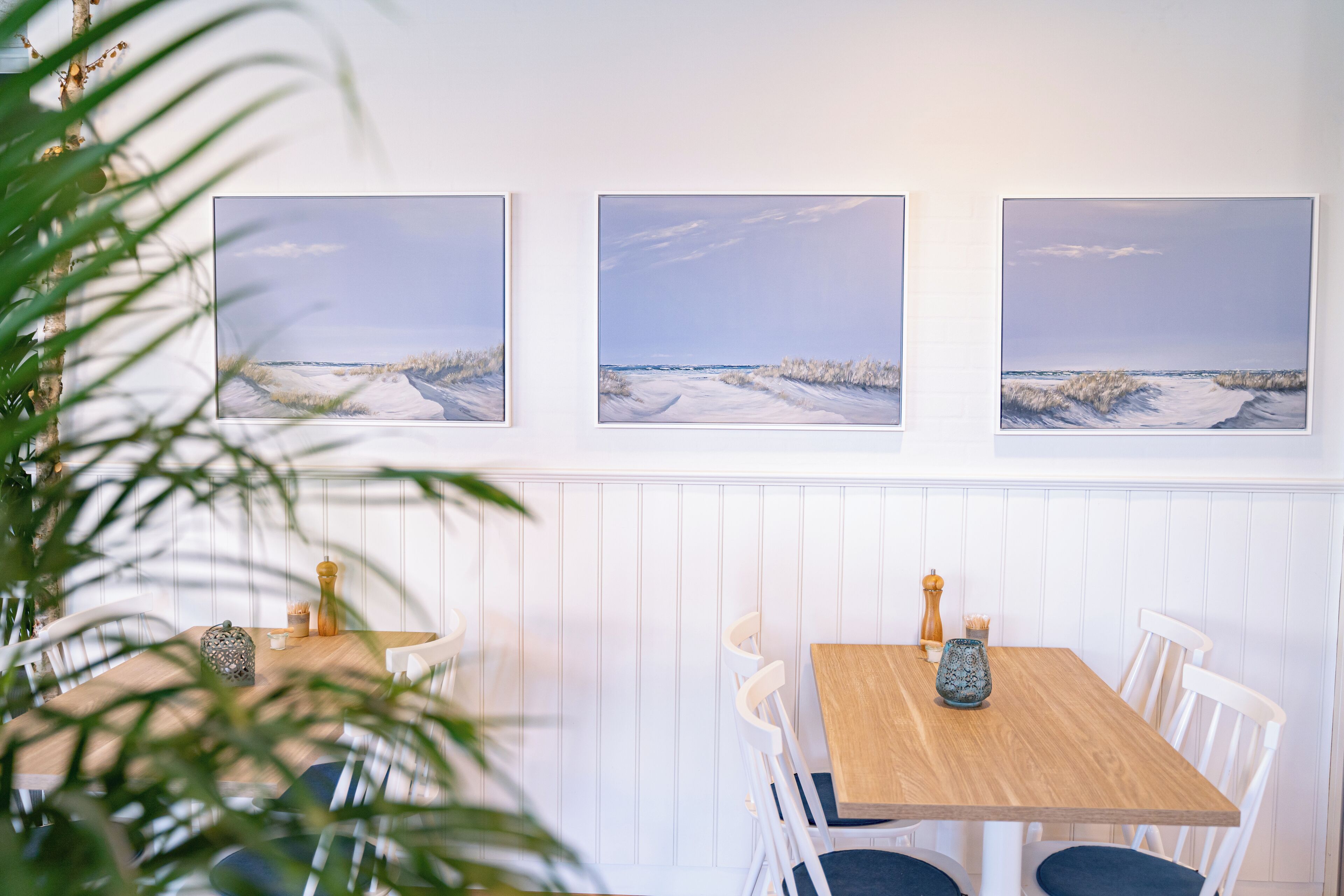 A dining area with a wooden table and white chairs, framed artwork above, and a plant in the foreground.