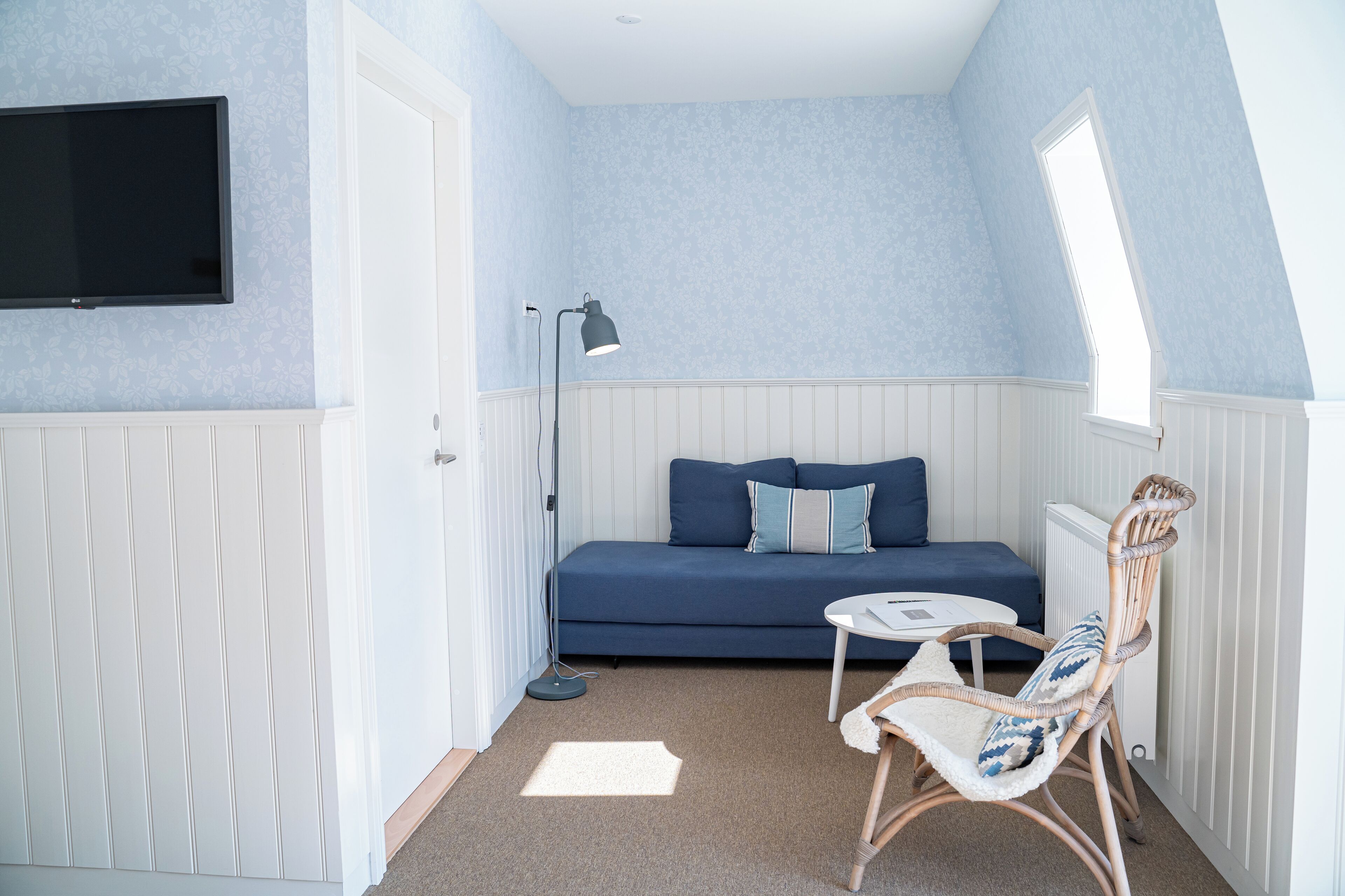 Una habitación pequeña y acogedora con un sofá azul, un sillón de mimbre, una mesita, una lámpara de pie y un televisor montado en la pared.