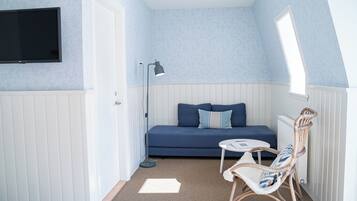 Una habitación pequeña y acogedora con un sofá azul, un sillón de mimbre, una mesita, una lámpara de pie y un televisor montado en la pared.