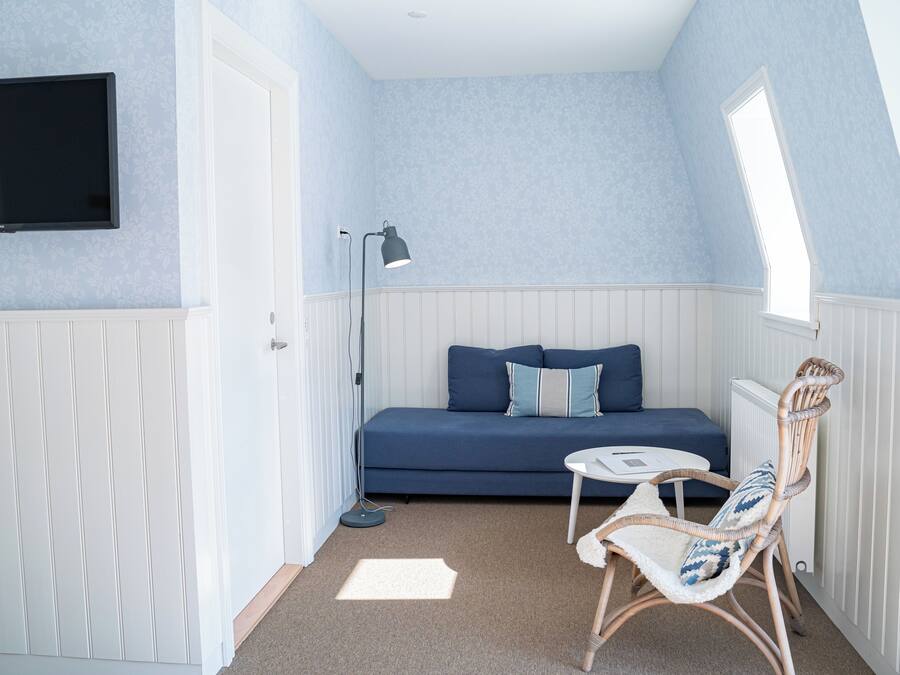 Una habitación pequeña y acogedora con un sofá azul, un sillón de mimbre, una mesita, una lámpara de pie y un televisor montado en la pared.