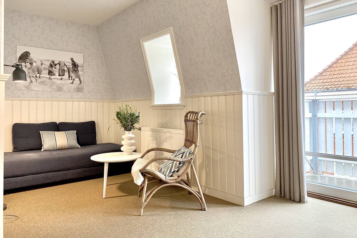 Una sala de estar moderna con un sofá oscuro, una mesita de noche blanca, una silla de mimbre, un espejo grande y vistas de un edificio a través de una ventana.