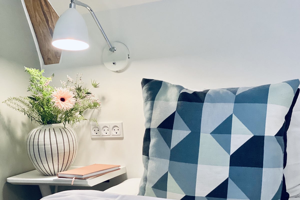 Habitación de hotel impecable con una cama, una mesita de noche con lámpara, un jarrón con flores y un cojín de estampado geométrico.