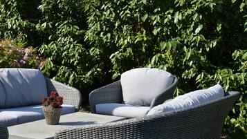 An outdoor seating area with wicker furniture and a concrete table, surrounded by lush greenery and a residential building with red-tiled roofs in the background.