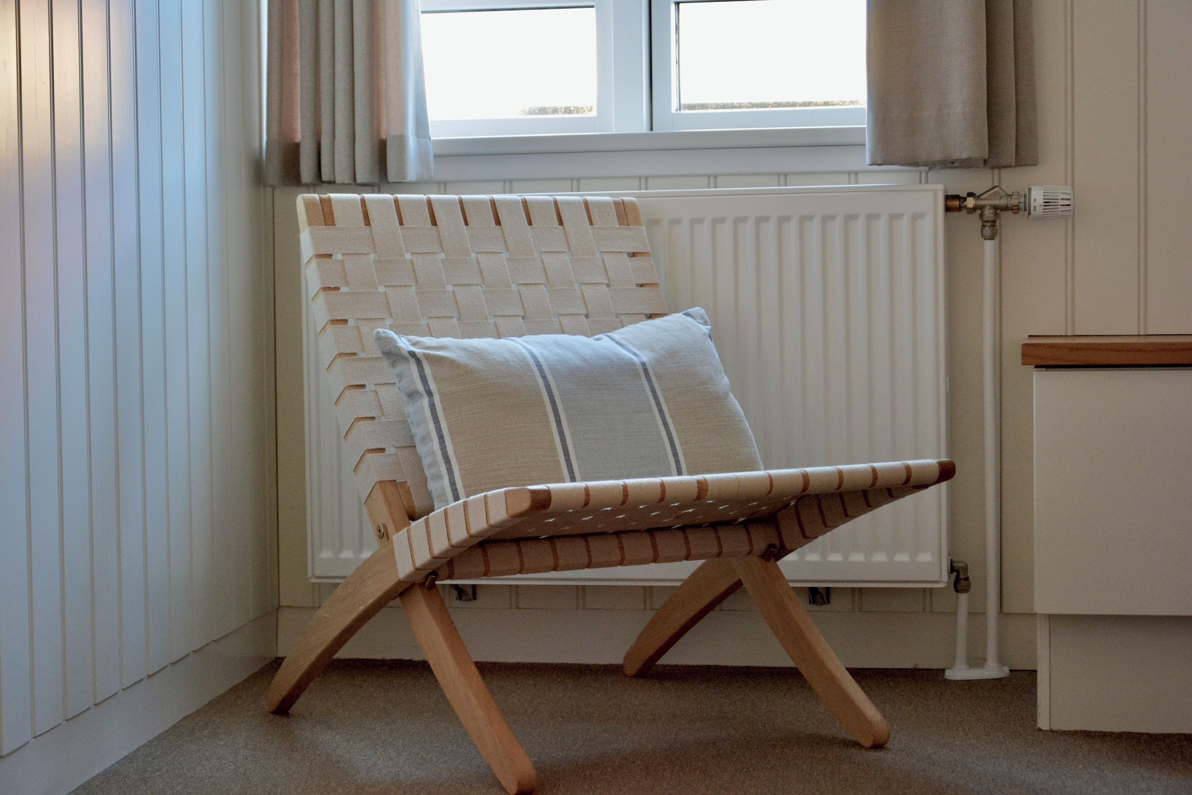 Ein Eckzimmer mit einem weißen Heizkörper, einem Holzstuhl mit einem gewebten Rücken und einem Fenster mit Vorhängen.