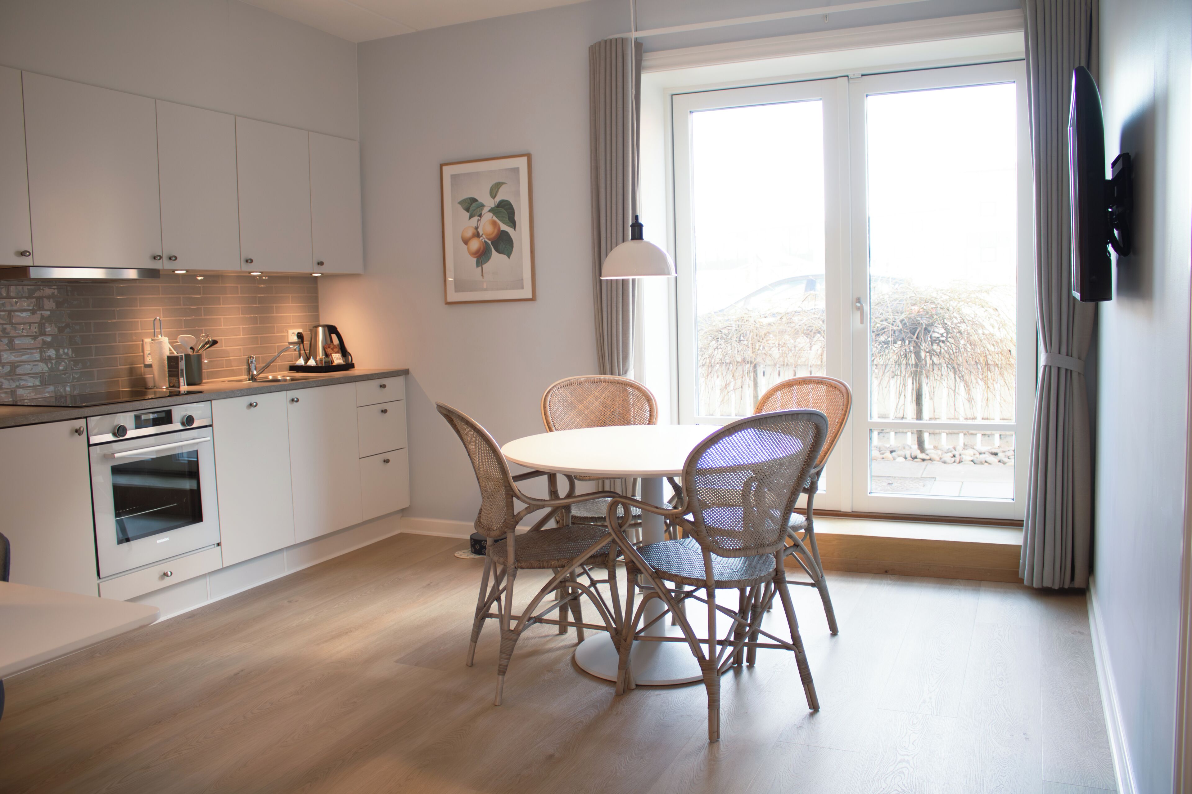 Eine moderne Küche mit weißen Schränken, einem eingebauten Backofen, einem Esstisch mit Korbstühlen und einem großen Fenster mit Vorhängen.