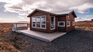 A wooden cabin with large windows, a deck, and a gravel ground. - Blue View Cabin 6A with Hot Tub (Bláskógabyggd)