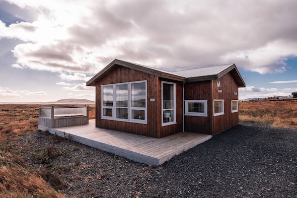 Une cabane en bois dotée de grandes fenêtres, d’une terrasse et d’un sol en gravier.
