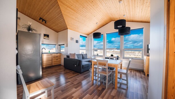 A modern kitchen with wooden flooring, a dining area with a wooden table and chairs, a sofa, and large windows offering a view of the outdoors. - Blue View Cabin 6A with Hot Tub (Bláskógabyggd)