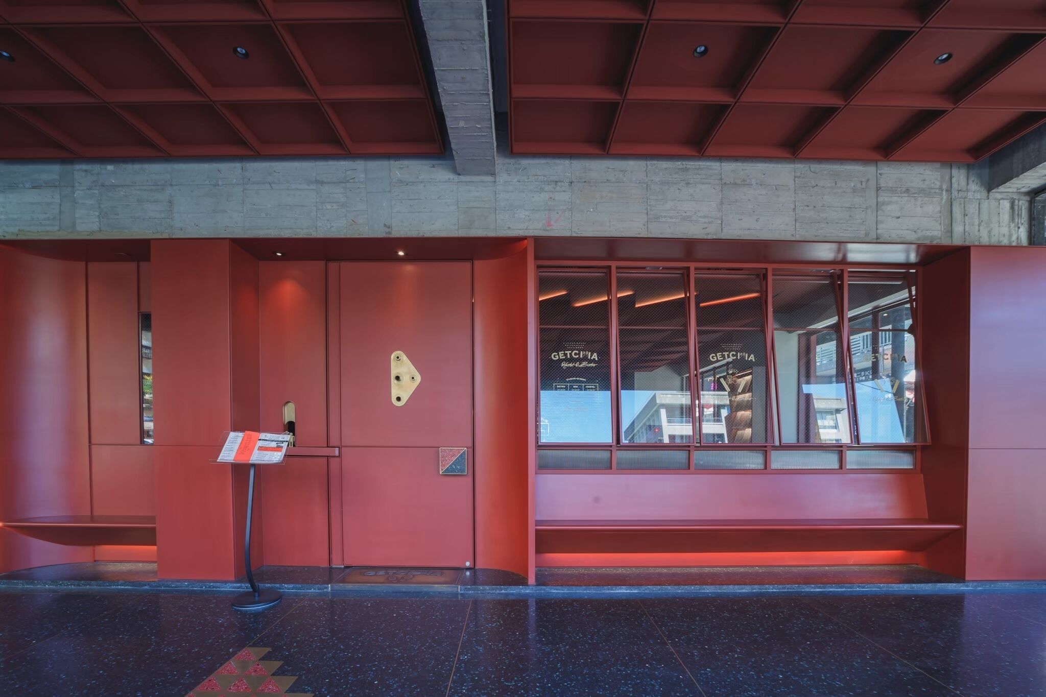 A modern building entrance with a red facade, a glass window displaying a sign, and a metal signboard.