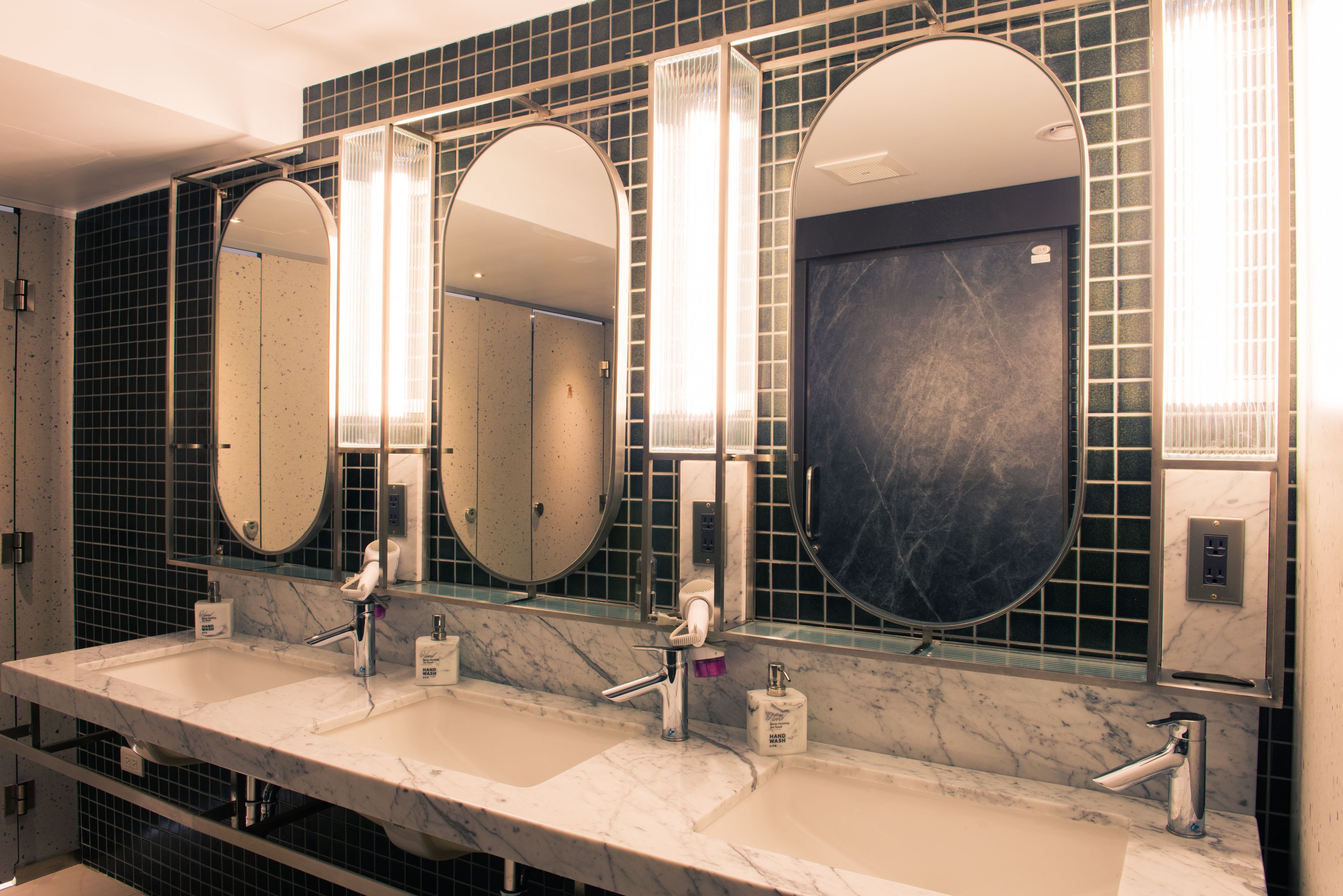 Un baño con tres lavamanos, cada uno con un espejo encima. Las paredes están cubiertas de azulejos con un diseño oscuro, y hay dispensadores de jabón en la encimera.