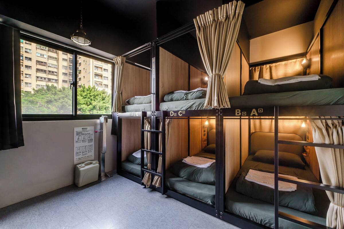 Quarto de hotel pequeno com beliches, uma janela com cortinas, abajur e uma mesa.