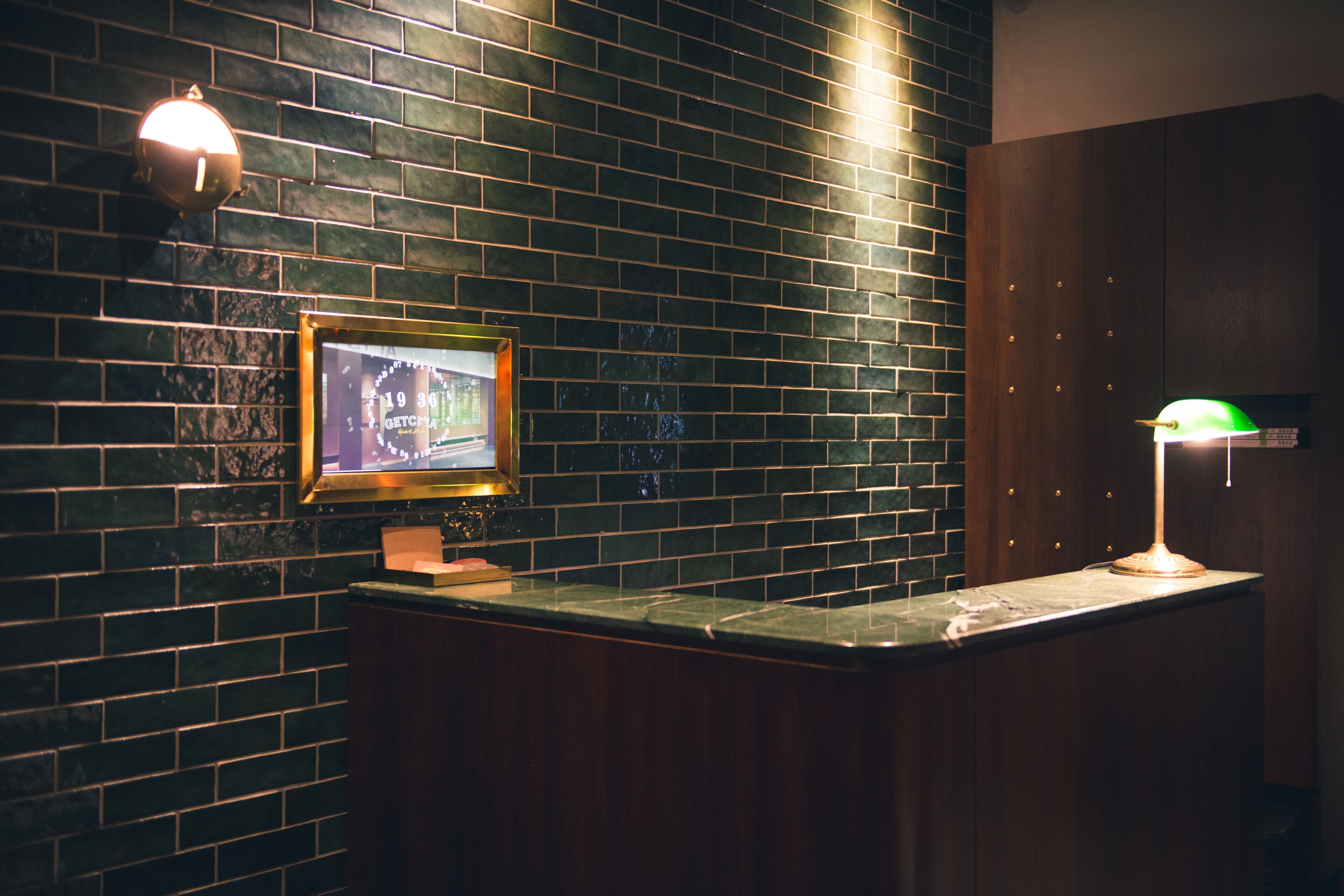 A reception area with a dark brick wall, a framed picture, a counter, and a green desk lamp.